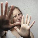 Portrait of a fearful woman in a gray tank top with hands pushed forward against a gray background.
