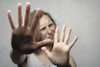 Portrait of a fearful woman in a gray tank top with hands pushed forward against a gray background.