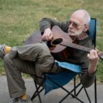 Elderly man seated outdoors playing acoustic guitar on a camping chair.