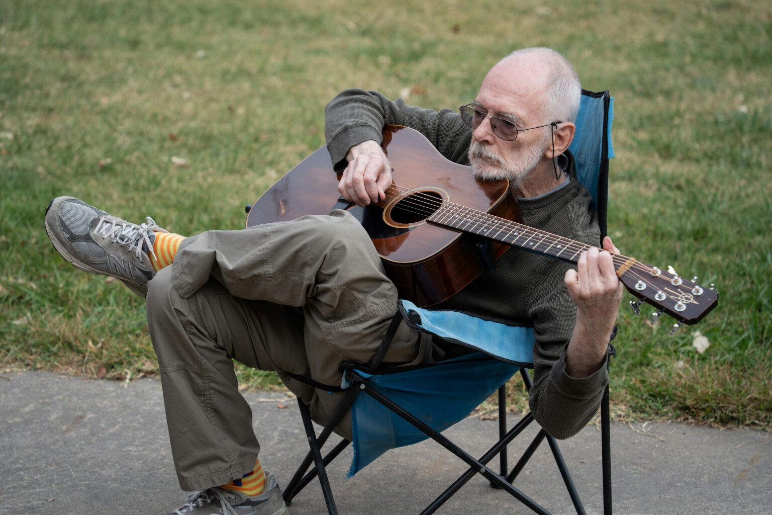 Elderly man seated outdoors playing acoustic guitar on a camping chair.