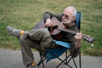Elderly man seated outdoors playing acoustic guitar on a camping chair.