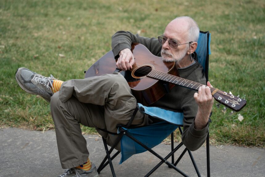 Elderly man seated outdoors playing acoustic guitar on a camping chair.