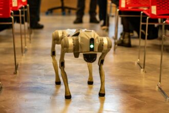 A robotic dog navigates an indoor setting amidst red chairs, showcasing technology in modern environments.