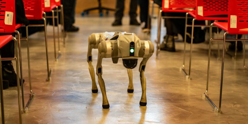 A robotic dog navigates an indoor setting amidst red chairs, showcasing technology in modern environments.