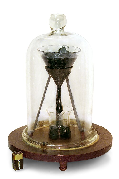 A glass funnel holding thick black pitch at the University of Queensland's Pitch Drop Experiment, with a 9-volt battery shown beside it for scale